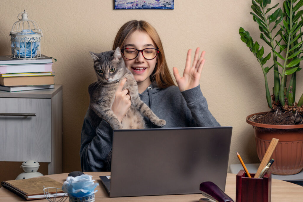 beautiful teenage girl with a cat in her arms sits in front of laptops. Teenage girl waves her hand at the laptop screen. Remote communication concept with friends or relatives